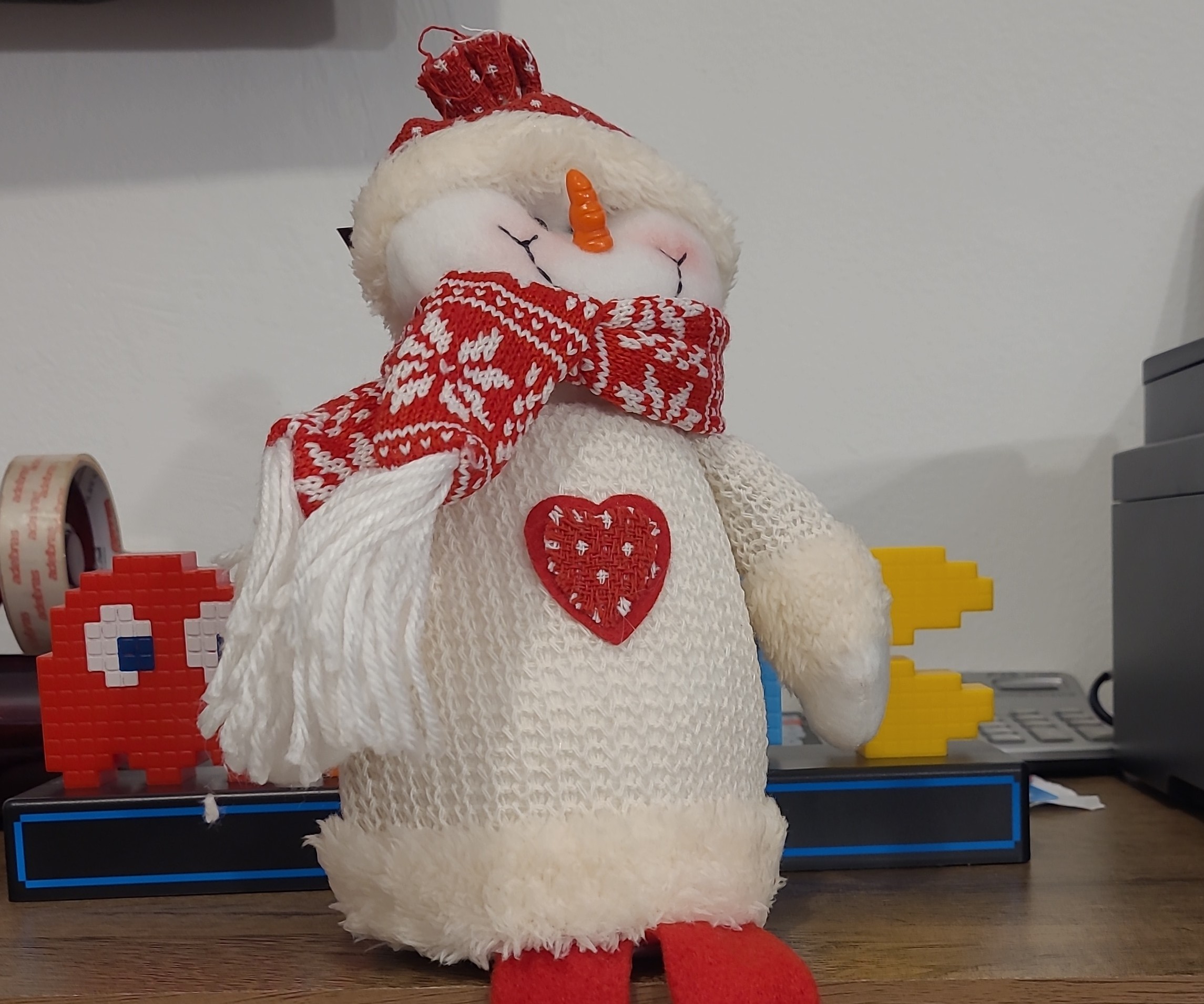 Descrição por Be My AI: Boneco de neve de pelúcia. Ele está usando um gorro vermelho com detalhe branco, cachecol vermelho e branco, além de uma decoração em formato de coração vermelho com detalhes brancos no peito. O boneco tem nariz de cenoura e olhos fechados feitos com linha preta, transmitindo expressão sorridente. Atrás dele, há algumas peças de decoração feitas de blocos de montar em formato de personagens do jogo Pac-Man (um fantasma vermelho à esquerda e o Pac-Man amarelo à direita). Há também fitas adesivas no canto esquerdo e parte de uma impressora ou equipamento eletrônico no lado direito da imagem. O boneco está apoiado sobre uma mesa de madeira.